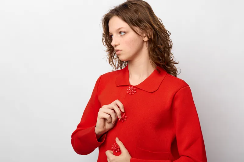 Young woman in red textured coat with decorative buttons looks sideways on white background. Fashion portrait with casual style and natural makeup in studio lighting.