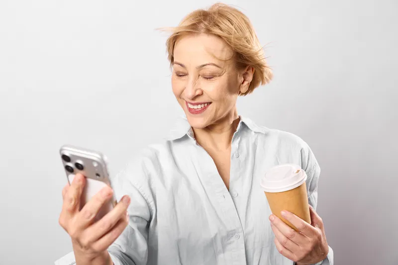 Senior woman smiling with smartphone, hands holding icecream, casual white shirt, soft gray background, communication moment, happy face indoors portrait