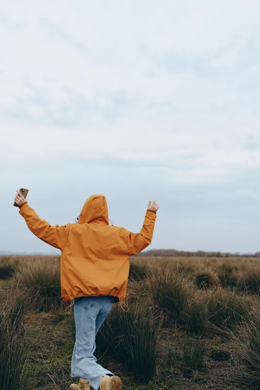 Woman lifestyle outdoors in a wide field, orange jacket and jeans, arms raised in joy, hood up against cloudy sky, standing on grass with soft wind, natural colors today