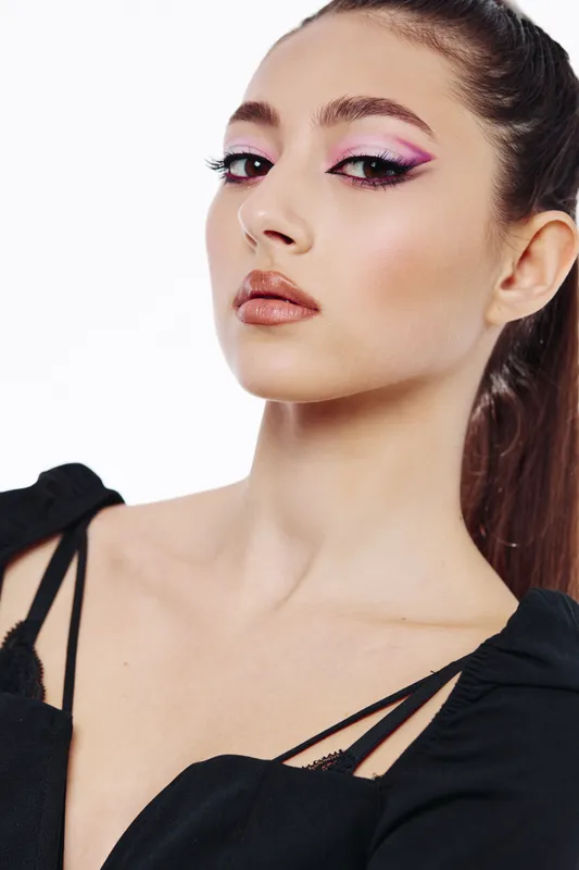 Young woman with creative pink eye makeup and glossy lips poses dramatically in a black top. High-fashion style for beauty and cosmetic concepts on white background.
