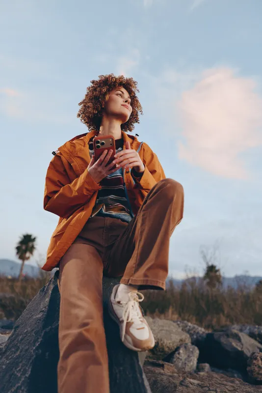 Woman outdoors taking a selfie with smartphone, curly hair visible, wearing a brown jacket, standing on rocks in nature, lifestyle travel mood under soft sky, warm evening light