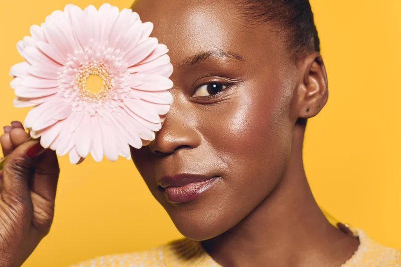 Close-up of a happy African woman with dark skin holding a pink flower near her face. Bright yellow background emphasizes natural beauty and floral concept for skincare and wellness.