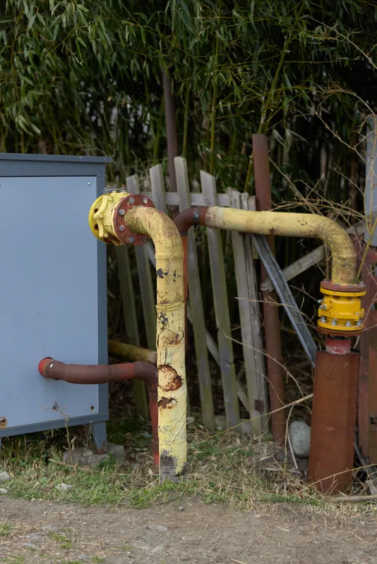 Outdoor garden water pipe valve with rusty metal, connected hose and utility equipment beside fence, weathered texture, sunlit yard ground, maintenance mood, natural greenery background, home