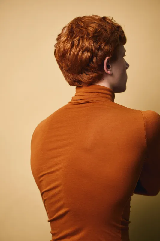 Redhead man in orange turtleneck sweater stands with back turned against beige background. Closeup of upper body and curly hair showing textured fabric and hairstyle details in studio light.