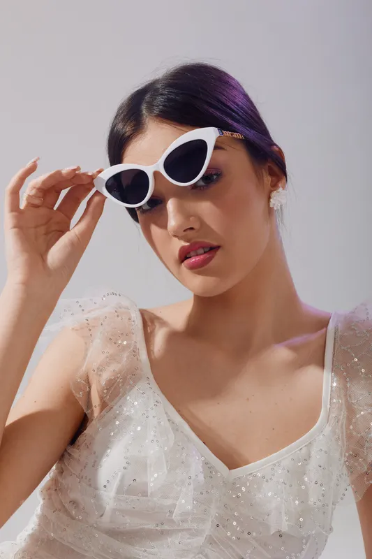 Young woman in a white elegant dress adjusts white sunglasses. Studio shot with soft lighting highlights her beauty and fashion style. Concept of glamour and summer accessories.