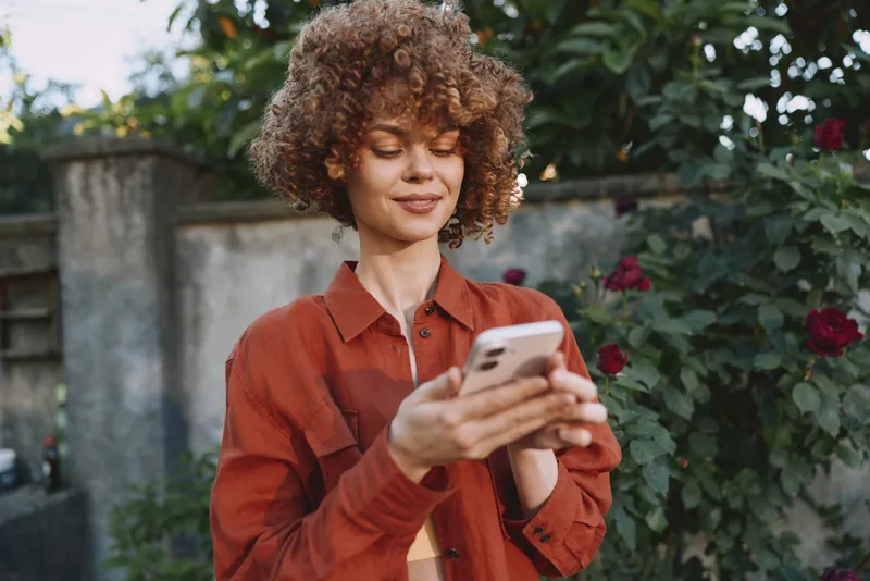 Young woman with curly hair checks her smartphone outdoors in a sunlit garden, wearing a rust jacket and a relaxed natural expression among blooming roses