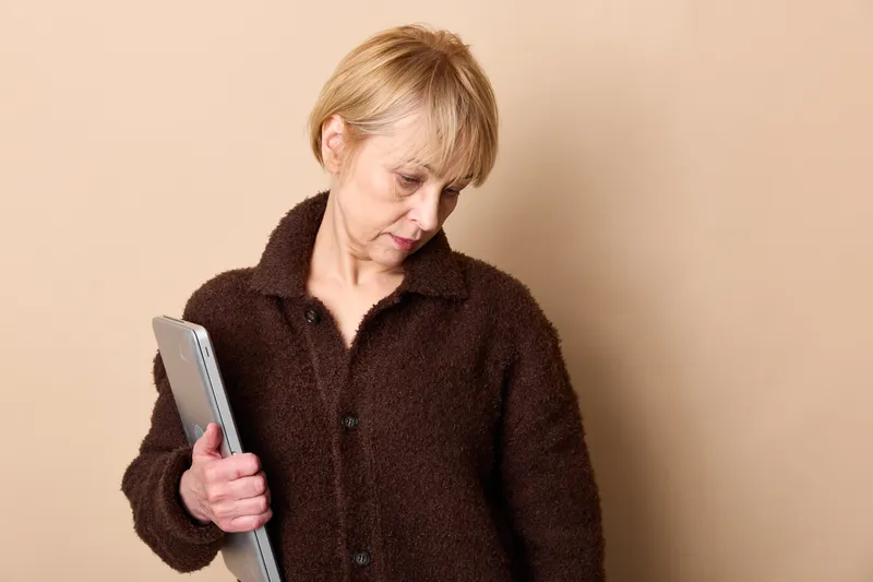 Elderly woman, worry in her expression, holding smartphone in hands, indoor beige background, communication tension, concept of anxiety, loneliness, stress, tense mood