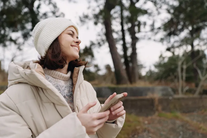 Woman in winter apparel outdoors, holding a phone and smiling, as trees and a fence backdrop create a candid moment of seasonal connectivity and relaxed nature
