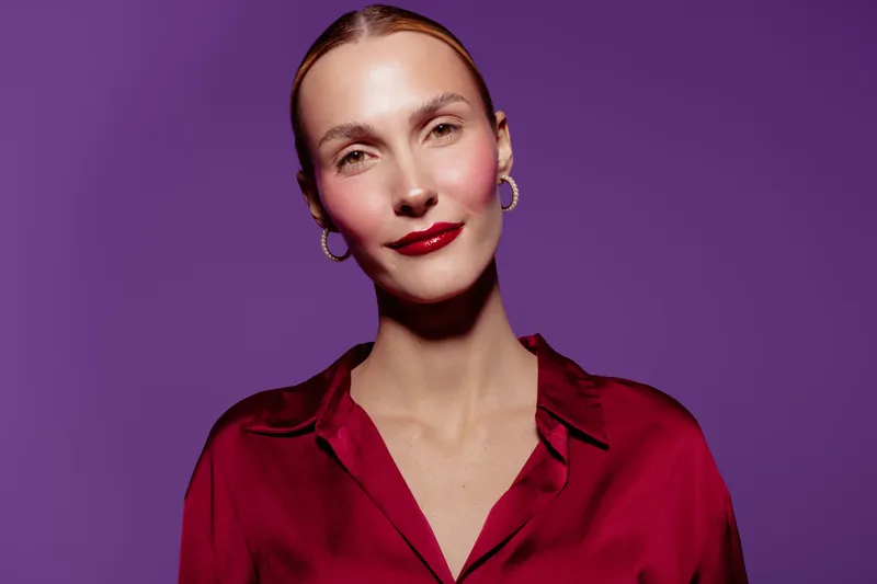 Confident woman with red lipstick and hoop earrings poses against purple background wearing red satin blouse. Concept of fashion, beauty, elegance, and modern style.