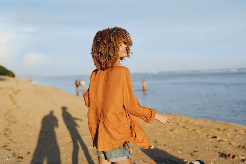 Girl at seaside by ocean beach in summer walking in sunlight, curly hair, child standing in denim shorts and orange top, looking at horizon, calm waves, breeze