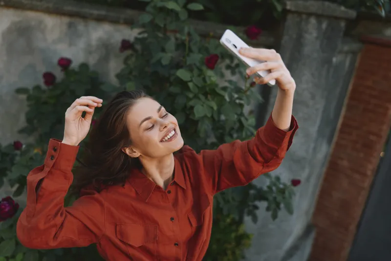 A cheerful woman in a red shirt captures a selfie outdoors, smiling at the camera in a sunny garden. Casual lifestyle moment focused on joy, spontaneity, and carefree everyday creativity
