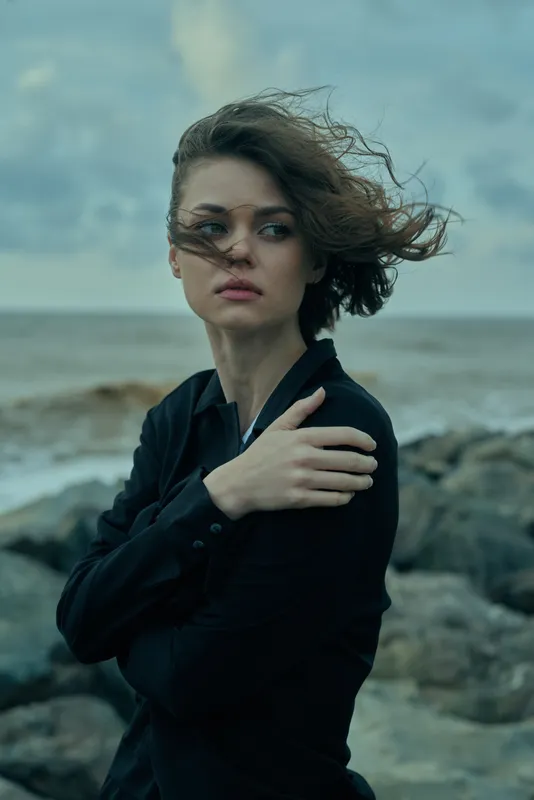 Woman on a windy coast, dark attire, hair swept by gusts, moody seaside scene with rocks and cloudy sky, expressive gaze, outdoor fashion photography, minimal color palette, natural light, candid pose