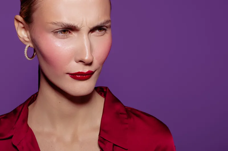Close-up of a serious woman with red lipstick and hoop earrings wearing a red blouse on purple background. Concept of fashion, beauty, makeup, and confident expression.