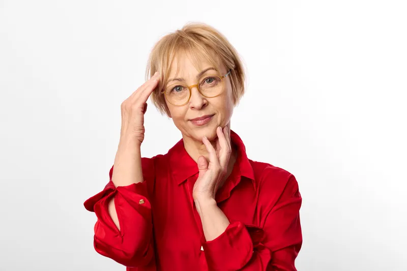 Woman senior with glasses in a red shirt, thoughtful portrait with stress, hand on face, isolated on a white background, calm studio expression, quiet concern now