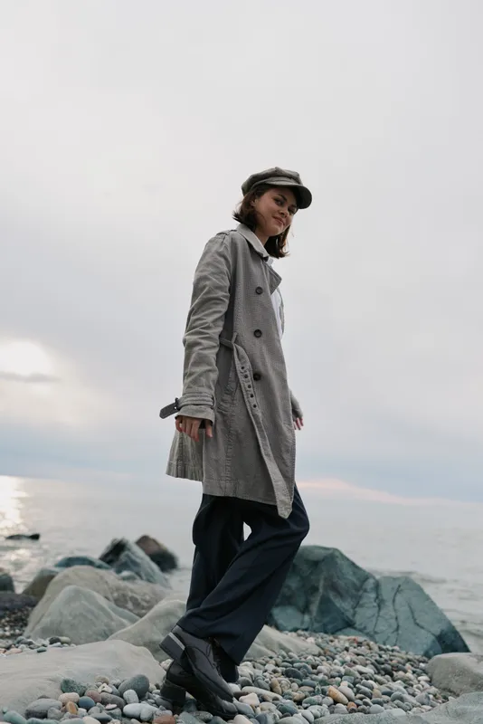 Young woman in long coat and cap walks on rocky beach near calm sea under cloudy sky. Casual fashion, outdoor lifestyle, and autumn concept with natural light and relaxed mood.