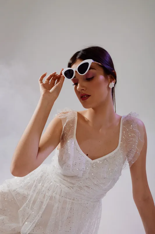 Elegant woman in a white sparkling dress adjusts white sunglasses. Soft lighting highlights her makeup and hairstyle. Concept of beauty, fashion, and glamour.