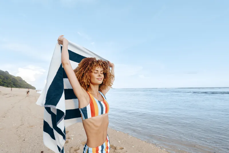 Smiling woman with curly hair holds a striped towel over her head on a sandy beach under bright sunshine, embodying summer leisure and carefree wellness moments