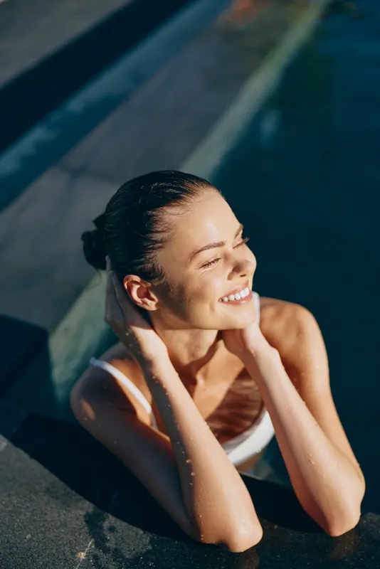 Young woman relaxes in outdoor pool with eyes closed and hands on neck enjoying sunny day in white swimsuit showing healthy skin and peaceful lifestyle moment.
