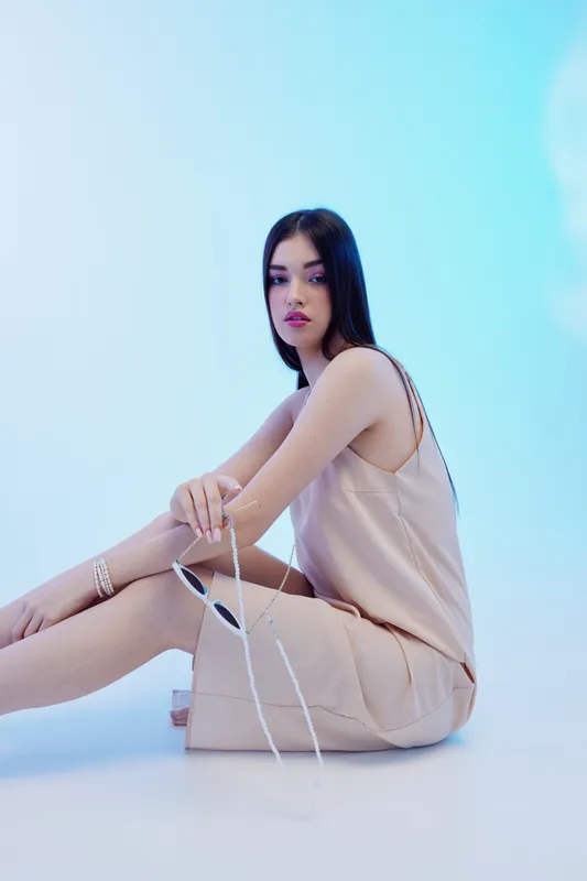 Young woman in beige dress poses sitting on floor with sunglasses in hand. Studio shot with soft blue lighting. Concept of beauty fashion style and modern elegance.