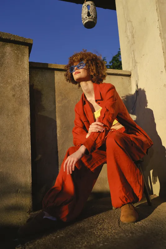 Woman lifestyle portrait wearing a red suit crouching on a sunlit terrace, stylish sunglasses, curly hair, bold fashion stance, architectural wall backdrop, urban chic mood