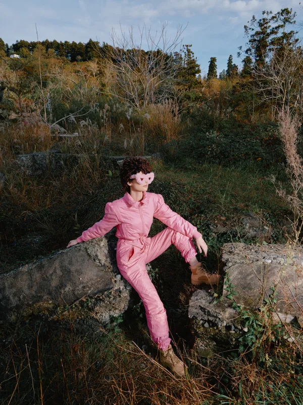 Doll in pink jumpsuit perched on a rock in a wild natural setting, surrounded by shrubs and autumn foliage, creating a whimsical outdoor portrait with a playful mood.
