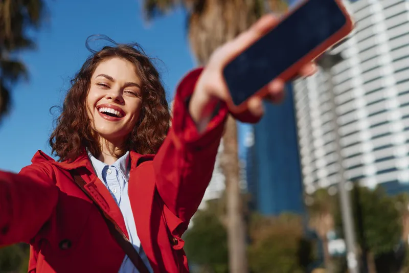 Cheerful woman outdoors, laughing, taking a selfie, sunny day, red jacket, casual pose, urban backdrop, palm trees, bright natural colors and sunshine