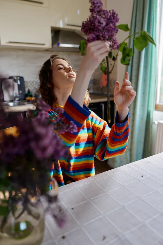 Woman in a colorful striped sweater arranges a bouquet indoors, showcasing home life, creativity and floral care in a bright kitchen with natural light and calm mood.