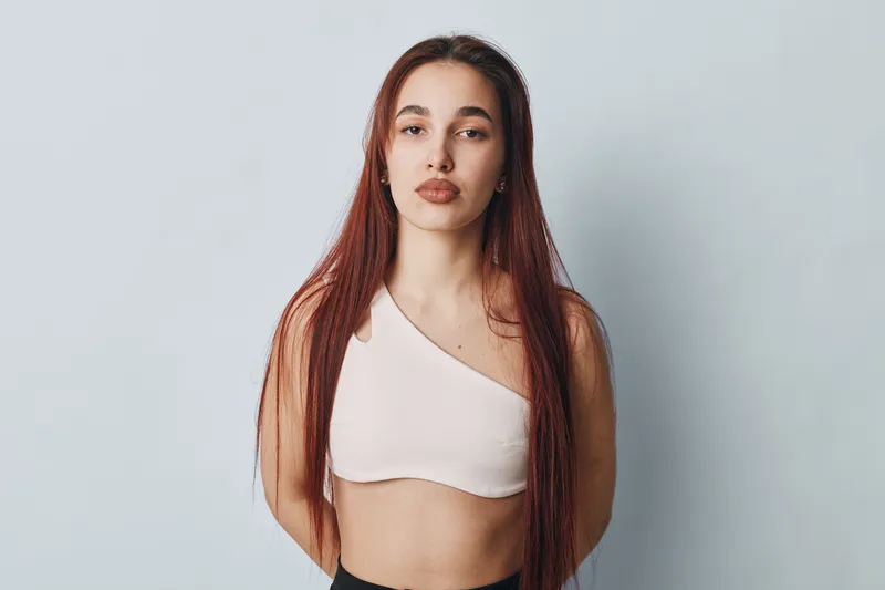 Young woman with long red hair wearing white one shoulder top standing against plain light background looking confidently at camera with neutral expression in studio portrait.