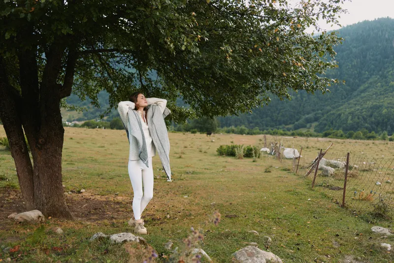 Joyful woman outdoors in a rural landscape, relaxed pose beneath a large tree, sunlit meadow backdrop, casual clothing, natural colors, serene moment