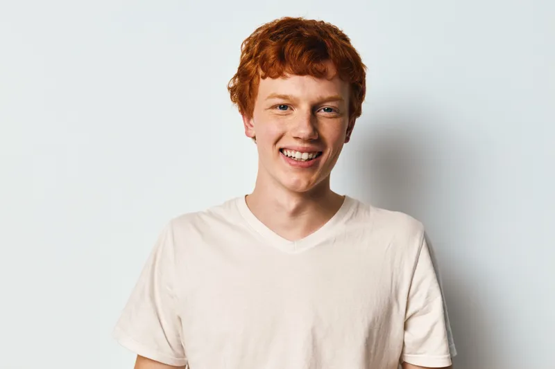 Young man with red hair smiling in white t shirt against plain background. Portrait of happy male person with freckles showing positive emotion and casual style.