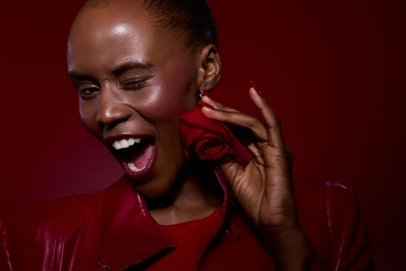 Portrait of a joyful dark skinned African woman winking and holding a red rose near her face. She wears a red jacket and smiles brightly against a dark red background. Concept of happiness.