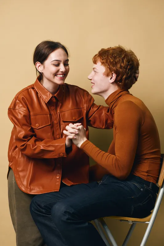 Young couple sitting and holding hands, smiling at each other in casual warm clothing, showing affection and connection in intimate friendly moment indoors.