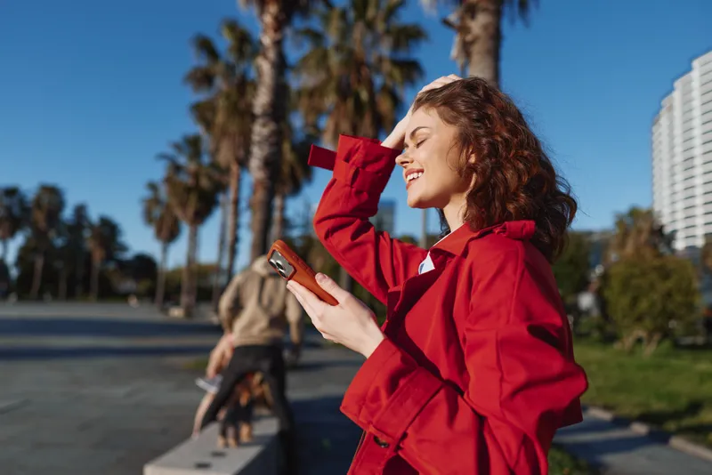 Smiling woman with curly hair wearing red jacket holding smartphone outdoors on sunny day. Urban park background with palm trees and modern buildings. Casual lifestyle concept.