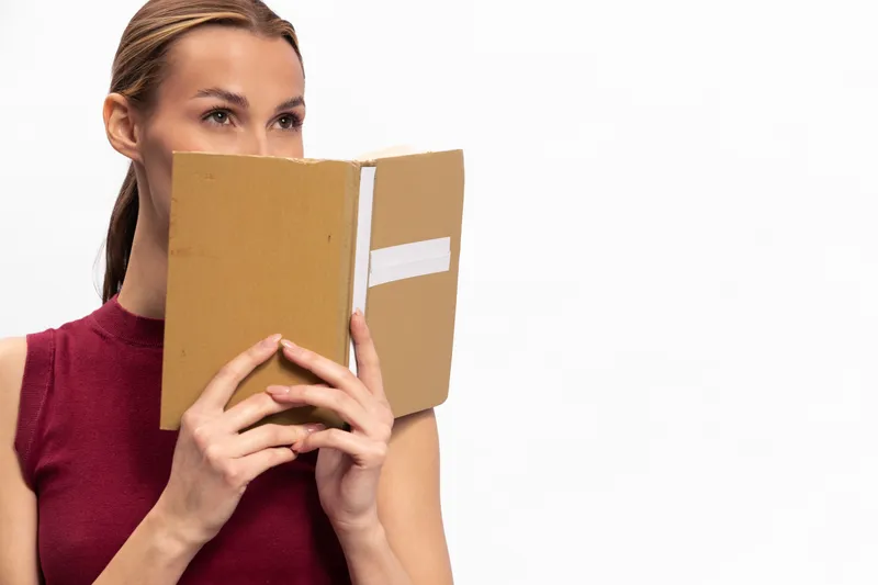Young woman with light hair holds a brown notebook in front of her face, wearing a sleeveless red top. Concept of privacy, study, and contemplation on white background.