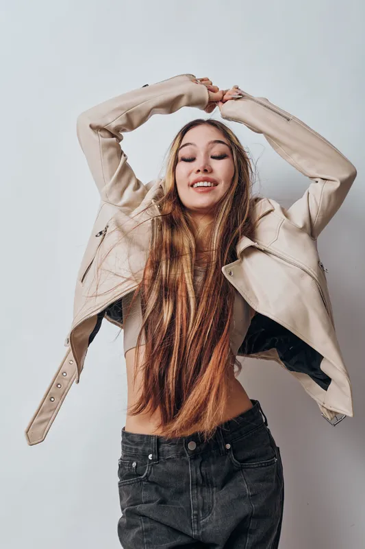 Young woman dancing with closed eyes wearing casual leather jacket and black jeans, long hair and smiling on light background expressing freedom and joy.