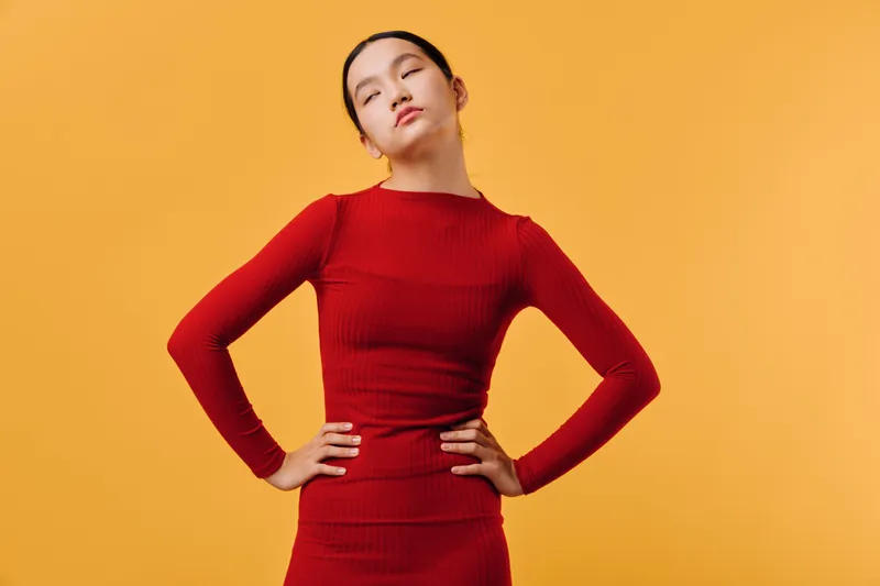 Young woman in red dress posing with hands on hips and eyes closed against orange background, showing confidence and style in studio lighting portrait of fashion model.