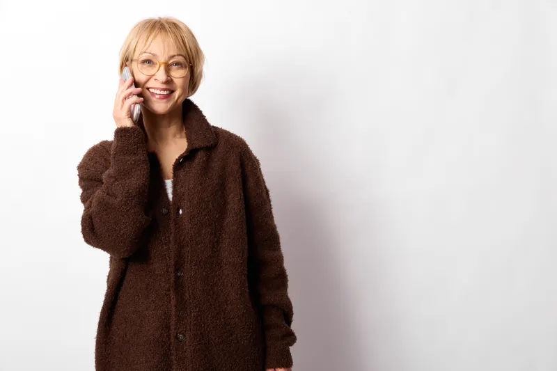 Smiling woman with glasses in a brown coat holding a phone to her ear, facing camera. Clean white background, indoor portrait. Communication call concept with a warm, calm mood.