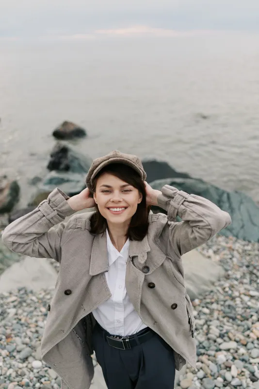 Smiling young woman in a hat and patterned coat enjoys a pebble beach with calm water in the background. Casual autumn fashion and relaxed outdoor lifestyle concept.