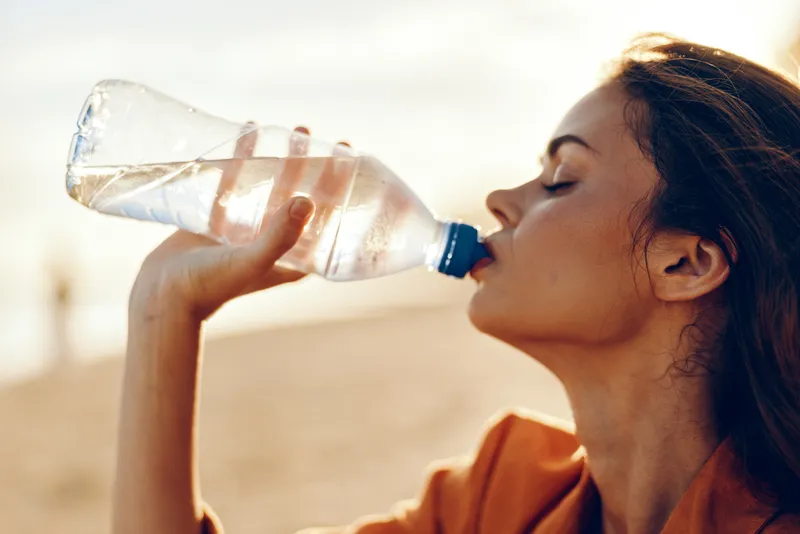 Young woman drinking water from a plastic bottle outdoors during sunset. Close-up side view with warm natural light. Concept of hydration, health, and refreshment in summer.