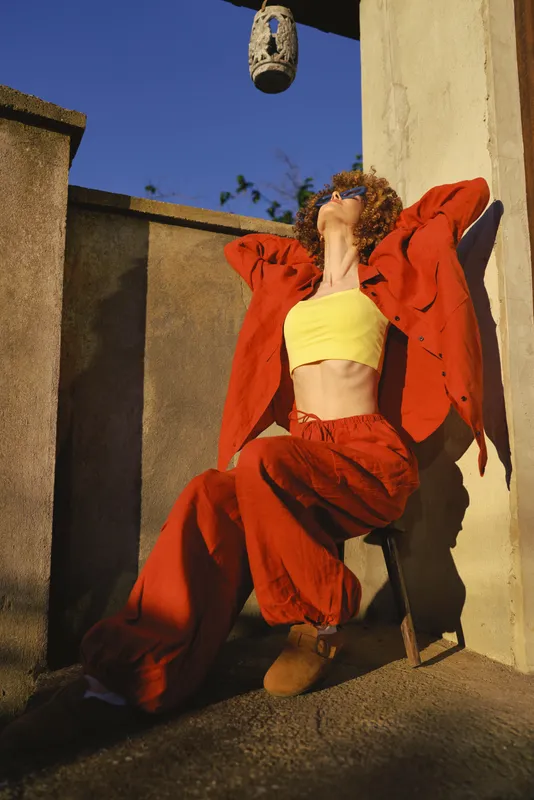 A woman in bold red jacket and yellow top leans back in an outdoor setting, capturing a striking lifestyle moment of confidence, movement, and casual fashion