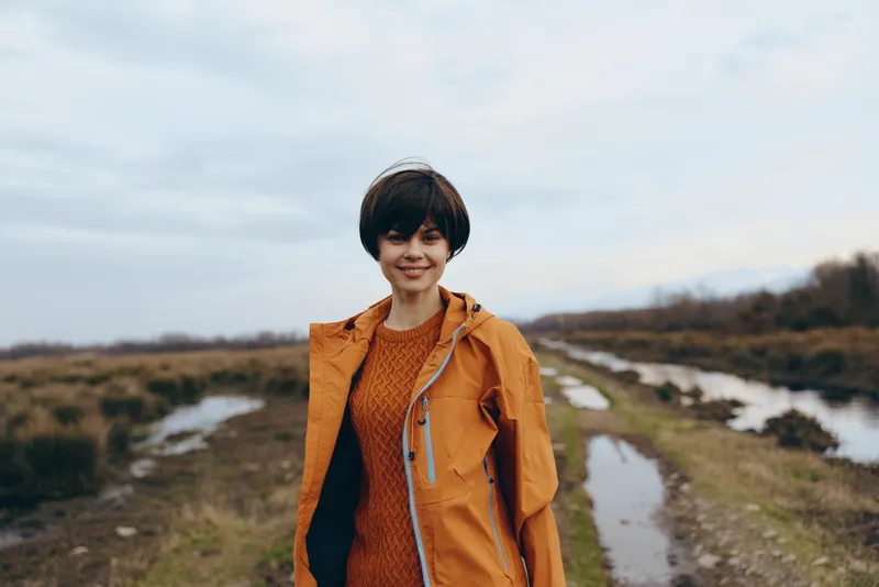 Smiling woman lifestyle outdoors on a riverside path wearing an orange coat and hat, walking near a winding river, calm nature scenery, soft daylight, travel mood during cool spring day