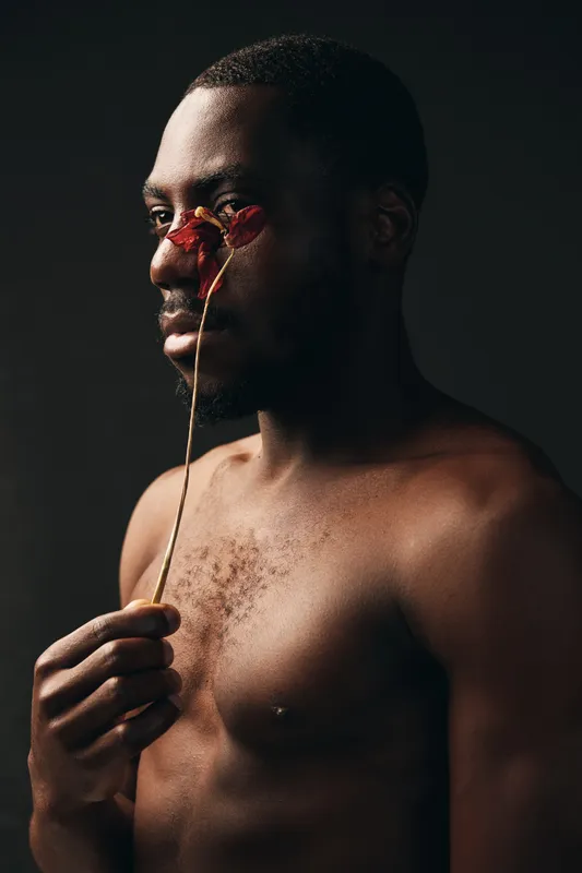 Creative conceptual portrait of a shirtless black man holding a dried flower near his face in a dark studio. Artistic visual style with emotional and thoughtful expression.