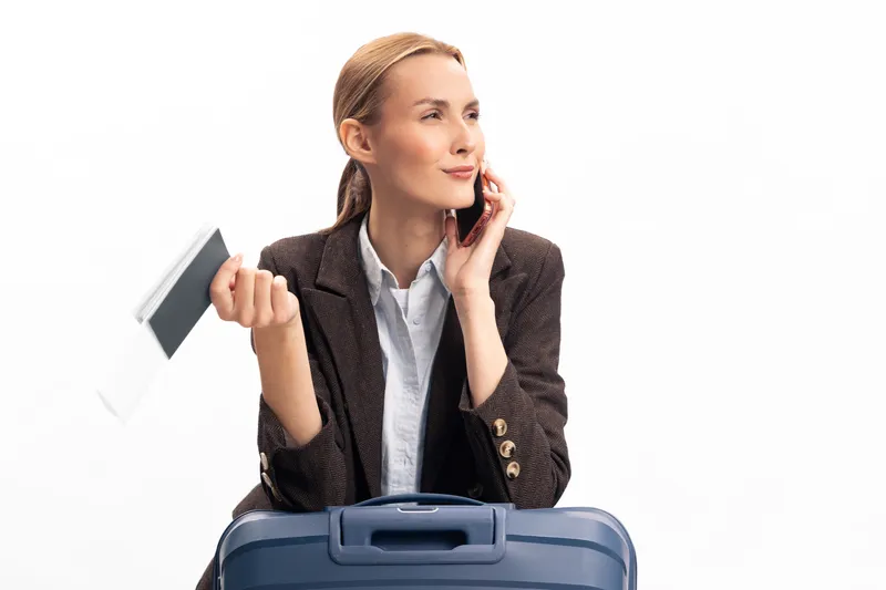 Businesswoman with suitcase holding passport and boarding pass talks on phone, preparing for travel. Concept of business travel, trip planning, and communication on white background.