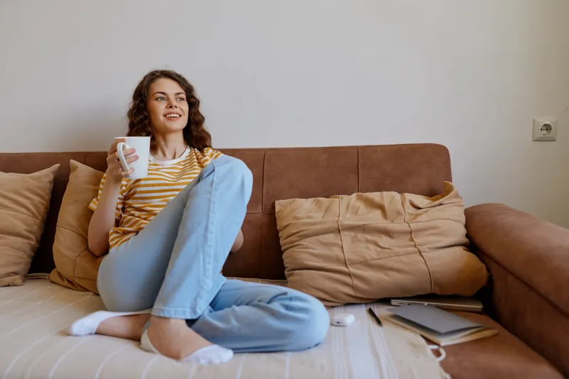 Cheerful woman home lifestyle on a couch, sipping from a mug, casual striped top, jeans, natural light, cozy living room, relaxed moment, candid and serene
