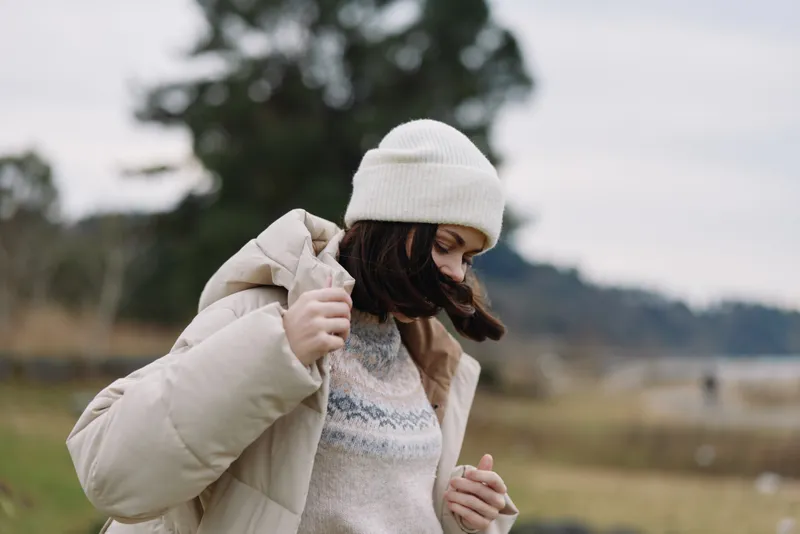 Winter outdoor scene with a woman wearing a warm coat and beanie, adjusting her jacket in a chilly landscape, soft natural light and distant trees creating a calm mood