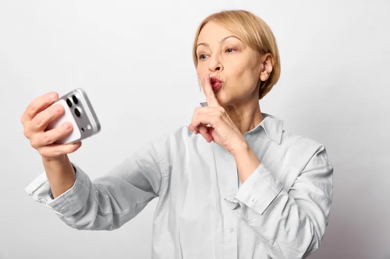 Surprised woman in light shirt holds a smartphone, finger to lips in a hush gesture, isolated on white background, concept of secrecy and instant shock.
