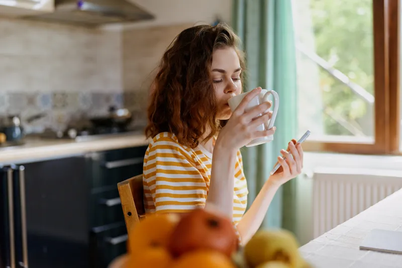 A woman enjoys a calm morning at home, sipping from a mug in a bright kitchen, with fruit on the table. A lifestyle scene focusing on comfort, routine, and mindful self care.