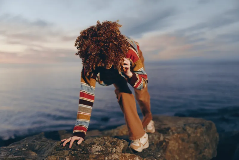 Woman lifestyle on an outdoor cliff by the sea at sunset, wearing a colorful sweater, bending forward in a candid pose, hands on rock, soft evening light with curly hair, calm expression