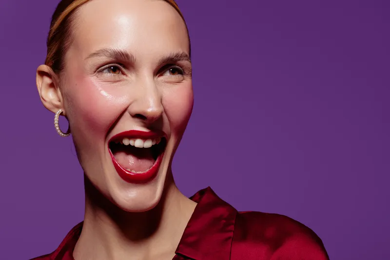 Happy woman with red lipstick and hoop earrings laughs joyfully against purple background. Close-up portrait with shiny skin and vibrant makeup creates energetic and positive mood.