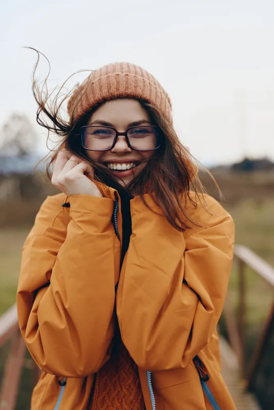 Smiling woman lifestyle outdoors in winter, wearing glasses and beanie, orange coat with collar held, wind moves hair, warm sunlight, blurred street background, casual fashion concept